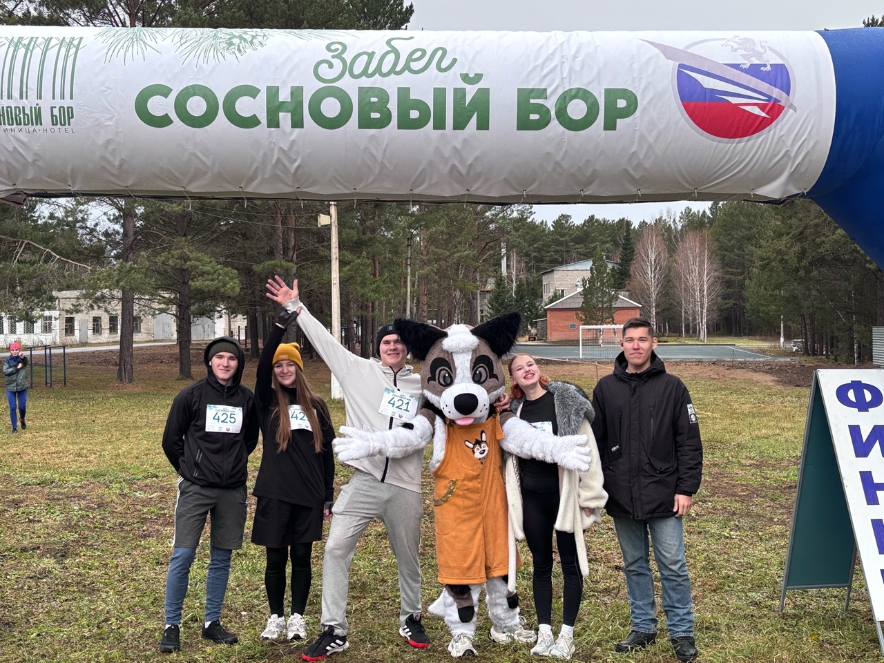 группа из шести человек стоит на открытом пространстве в лесу, рядом с большим надувным баннером с надписью "Забег сосновый бор". В центре внимания — человек в костюме собаки-маски. Участники обнимаются, смеются и делают позу для фотографии. Два человека в черной одежде, двое других — в ярких нарядах. Вдалеке виднеются деревья и здания. Атмосфера веселая, ритуал праздника и командного духа.