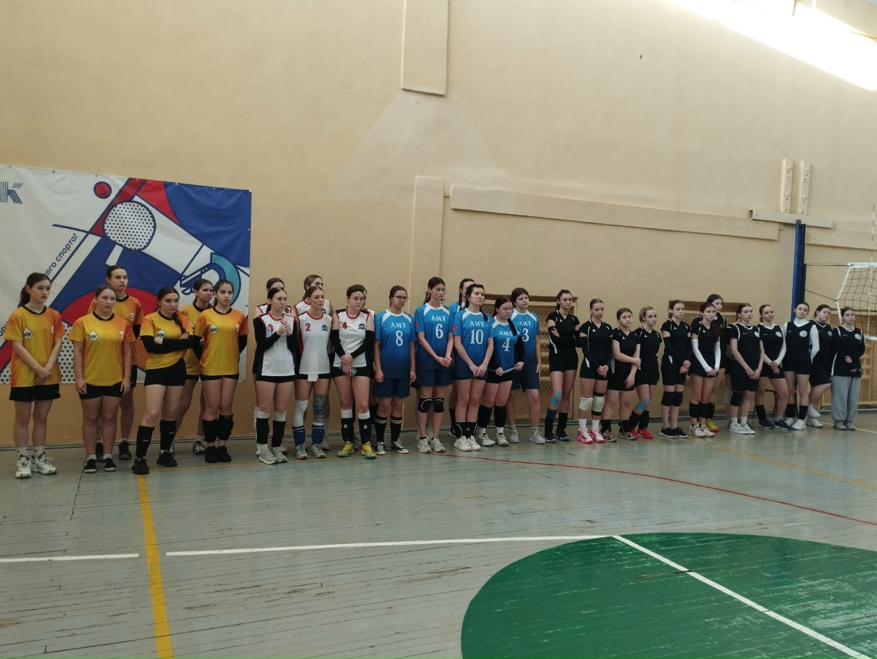 представлена группа девушек, стоящих в линию на волейбольной площадке. Команды находятся на турнире или соревновании. Каждая команда одета в свою цветовую форму: у одних девушек яркие желтые футболки, а у других — черные и белые. Фон содержит спортивные элементы, такие как мяч и сетка.