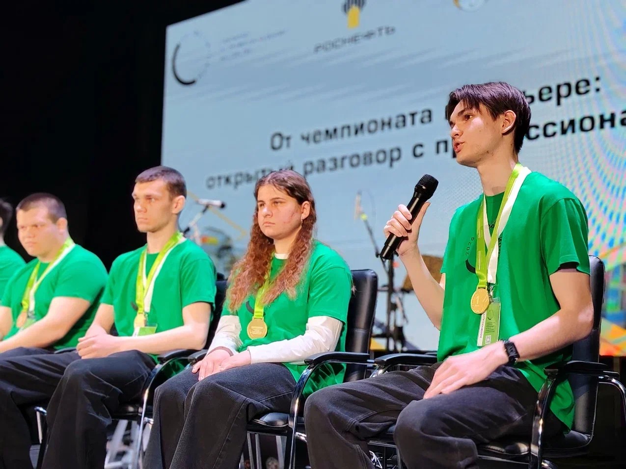 На сцене четыре молодые люди в зелёных футболках с медалями. Слева направо: два молодых человека, один с короткой стрижкой, другой с длинными волосами. Посередине девушка с длинными волосами, а справа молодой человек, который говорит в микрофон. Сзади на экране информация о чемпионате.