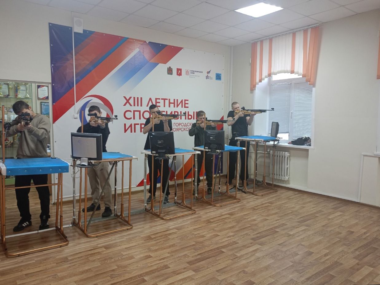 На изображении группа людей, занимающихся стендовой стрельбой. Все они стоят за столами с тренажерами для стрельбы, выглядят сосредоточенными. В центре стола находятся устройства для стрельбы, а вокруг — спортивные участники в различной одежде. Стена украшена баннером с логотипом спортивных игр, подчеркивая коллективный характер мероприятия.