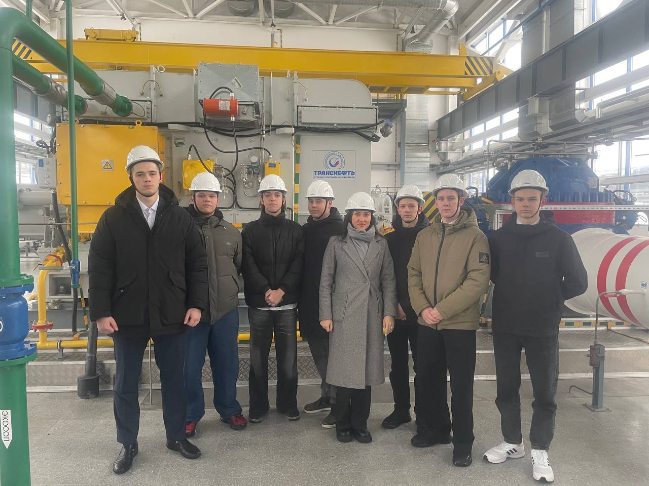 На фотографии группа из восьми человек, стоящих в помещении промышленного завода. Все они носят каски безопасности. На переднем плане несколько мужчин и одна женщина, одетые в тёплую одежду. Фон состоит из большого промышленного оборудования, включая трубы и серые стены. В воздухе ощущается ощущение работы и производственной активности.