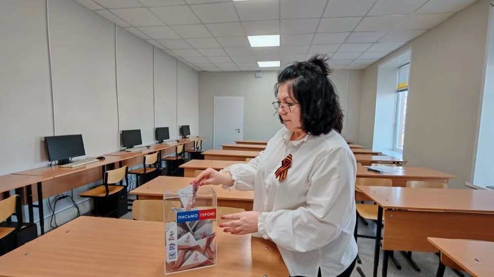 изображении женщина, возможно, преподаватель или организатор, стоит в учебном помещении с современным интерьером. Она одета в белую рубашку и держит прозрачную коробку с наклейками, на которых написано "Тест" и "Голосуйте". Вокруг неё - столы, на которых расположены компьютеры, указывая на то, что это класс для занятий или презентаций. Женщина сосредоточена и, похоже, готовит что-то, связанное с голосованием.