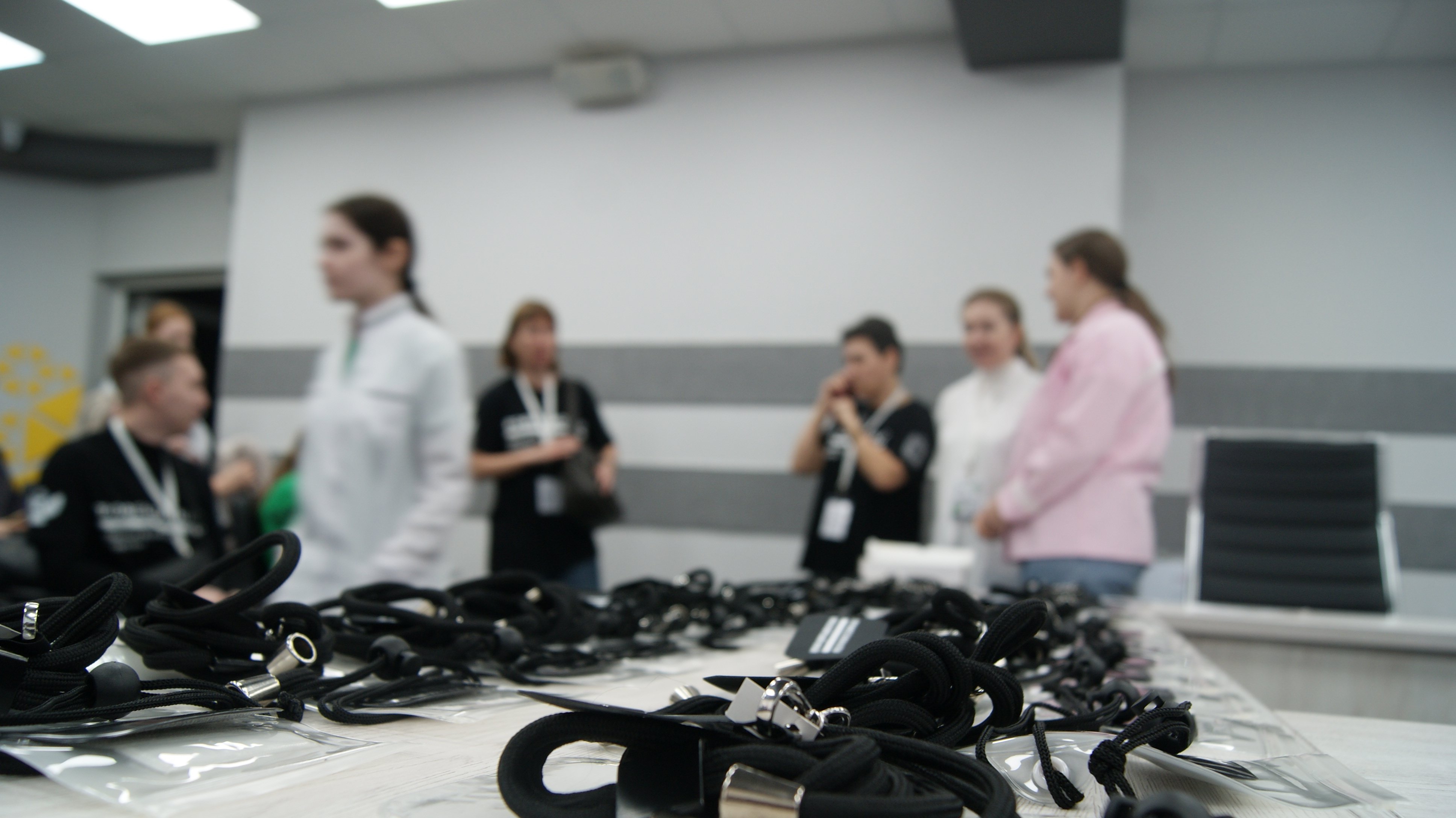 На этом изображении в фокусе множество черных шнуров, лежащих на столе. На заднем плане группа людей в белых халатах и обычной одежде обсуждает что-то между собой. Один из участников держит в руках пластиковый предмет, возможно, некий рабочий инструмент. Атмосфера выглядит непринужденно, все заняты общением.