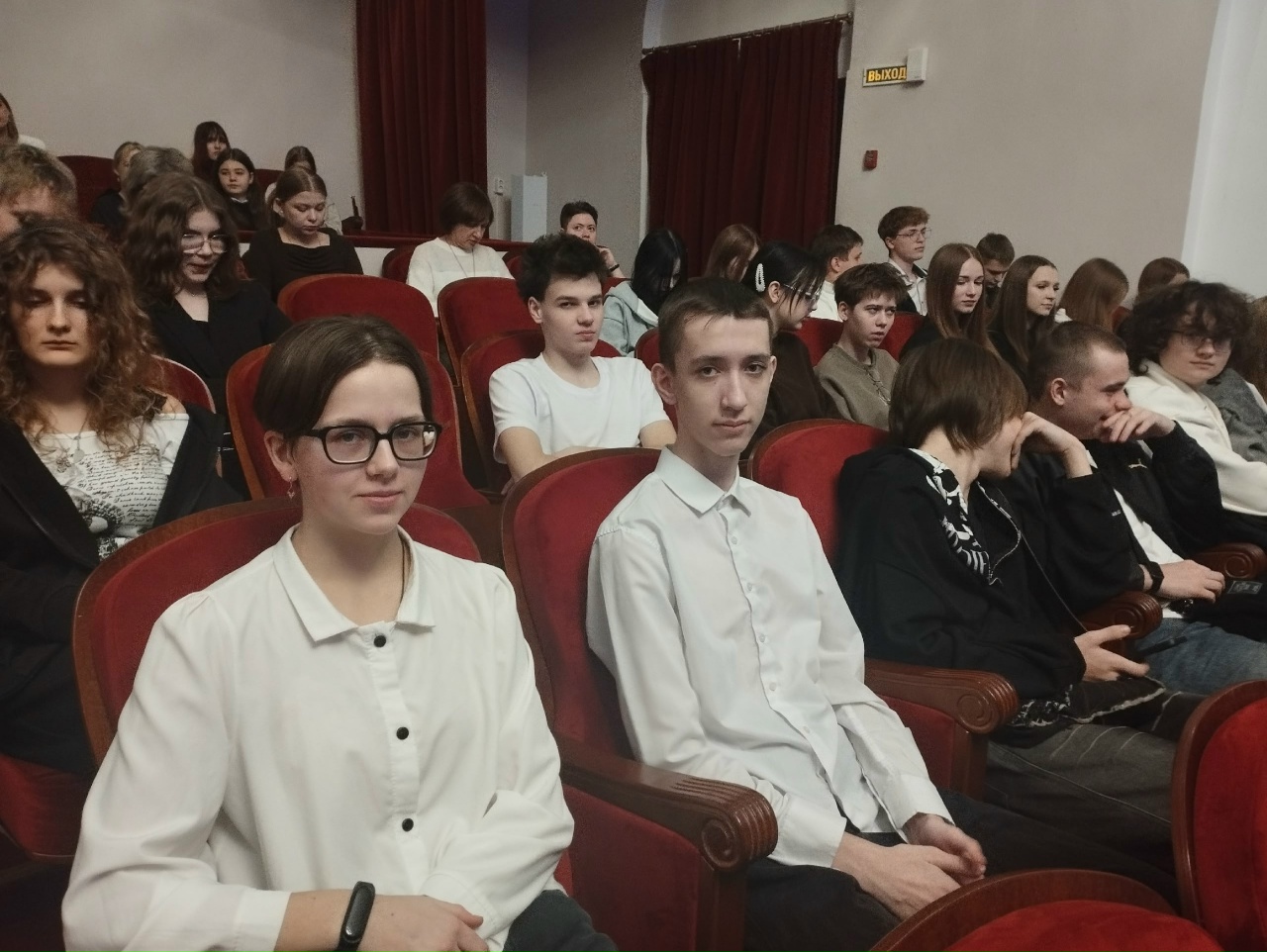 На этом снимке показан зал театра, заполненный зрителями. В центре изображения двое молодых людей: парень и девушка, сидящие рядом. Девушка в белой рубашке с короткими рукавами и большими очками. Она смотрит вперёд, в то время как парень в светлой рубашке также обращён в сторону сцены. На заднем плане видно множество зрителей, у которых разнообразные прически и одежда.