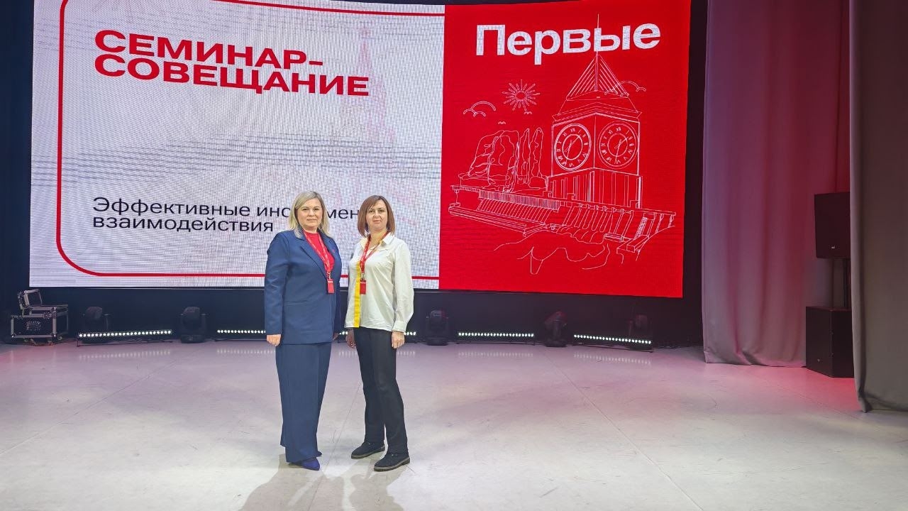 На фотографии изображены две женщины, стоящие на сцене. Обе одеты в деловые наряды. На заднем плане находится крупный экран с надписью «Семинар-совещание» и подзаголовком «Эффективные инструменты взаимодействия». Освещение сцене яркое, что создает атмосферу делового мероприятия.