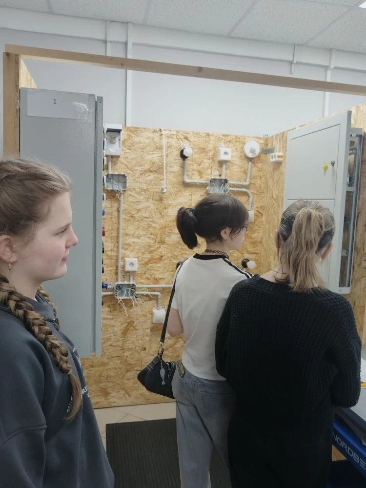 снимок фокусируется на трех молодых женщинах, стоящих перед стеной из дерева с электрооборудованием и проводами. Девушки внимательно смотрят на что-то перед собой, возможно, на демонстрационное устройство. Обстановка кажется учебной, с элементами, характерными для технического обучения.