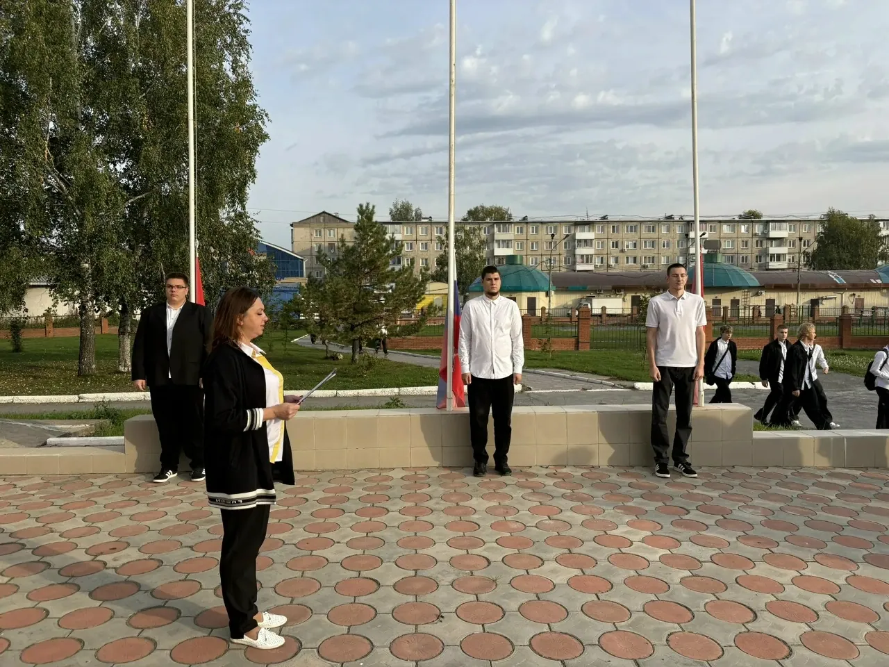 Женщина в черной одежде с белыми полосами держит бумаги и выступает с речью на мощеной площадке. Рядом с ней стоят молодые люди: один в черном костюме, двое других в белых рубашках и футболках стоят у флагштоков с флагами России и красным знаменем. На заднем плане видны жилые дома, деревья и проходящие мимо люди.