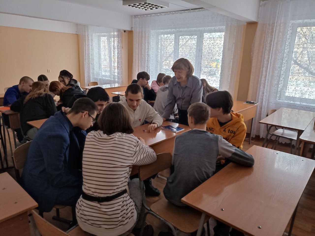 На этом снимке группа подростков сидит за столами в учебной аудитории. Учащиеся расположены в круге, обсуждая что-то с интересом. В центре внимания инструмент или учебный материал, на который все смотрят. Преподаватель, женщина среднего возраста, стоит у стола и направляет обсуждение. Светлые окна создают уютную атмосферу.