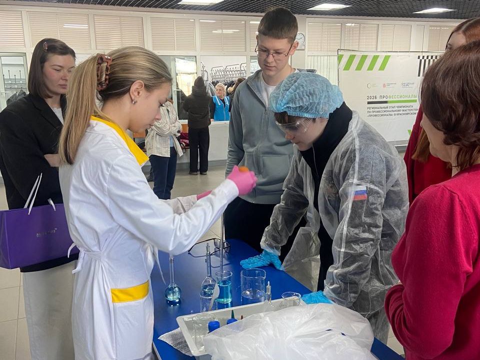 молодые люди принимают участие в эксперименте. Женщина в белом Lab-костюме и перчатках готовит реагенты в пробирках на синем столе. Два человека, стоящие рядом, внимательно наблюдают за процессом. Один из них одет в защитный халат и маску, а другой просто в рубашке и очках. На заднем плане видны другие участники мероприятия, которые наблюдают за процессом.