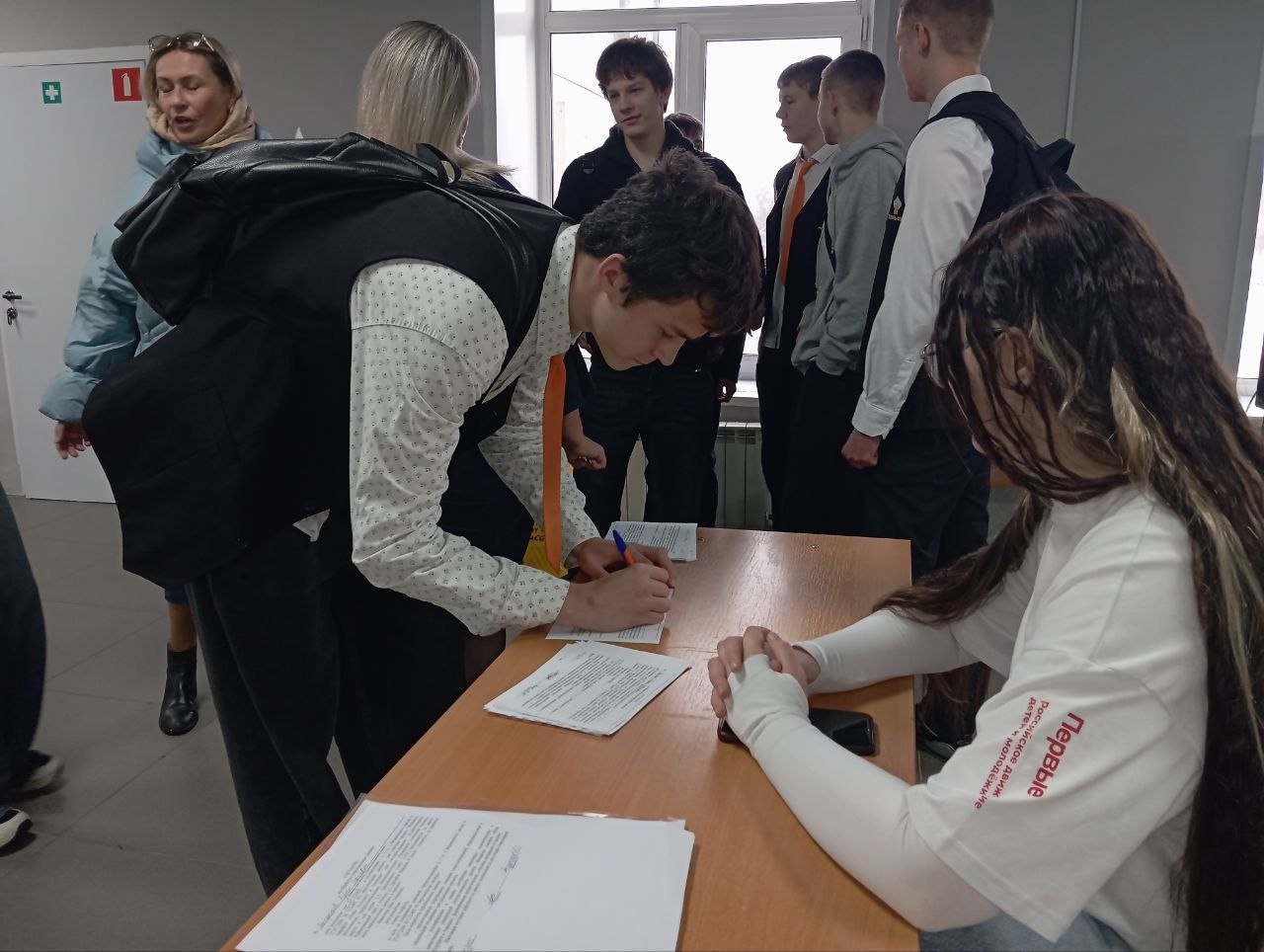 На фотографии показывается сцена в помещении, где несколько молодых людей взаимодействуют друг с другом. Один из парней стоит за столом и заполняет форму, в то время как девушка с длинными волосами сидит перед ним, скрестив руки. На заднем плане можно выделить других подростков, которые обсуждают что-то между собой. Обстановка деловая, с нейтральными цветами стен и мебели.