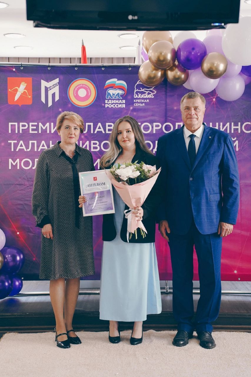 представлены три человека: две женщины и один мужчина. Одна из женщин держит букет цветов и диплом, что также свидетельствует о награждении. Фон украшен шарами и баннерами, создавая праздничную атмосферу.