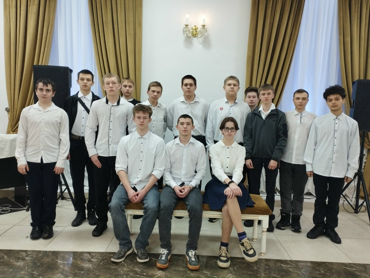 На фотографии группа молодых людей, состоящая из 15 человек, стоит в помещении с белыми стенами и яркими шторами. Все они одеты в белые сорочки. В центре группы — несколько человек, сидящих на скамье, большинство из них воздерживаются от выражения эмоций. Взгляды участников направлены вперед, а в заднем плане видны колонки и другие предметы, указывающие на то, что это мероприятие может быть связано с выступлением или концертом.