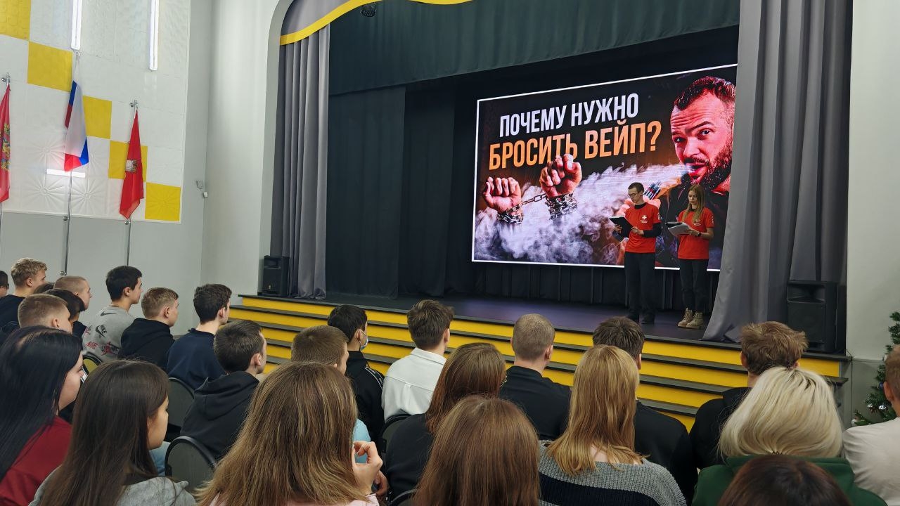 показана сцена с выступающими. На экране размещен заголовок с вопросом: "Почему нужно бросить вейп?" Лекторы держат микрофоны и обращаются к аудитории, которая снова состоит преимущественно из молодых людей. Зал оформлен флагами, освещение приглушенное, создавая уютную атмосферу для обсуждения темы. Зрители проявляют интерес, обращая внимание на экран.