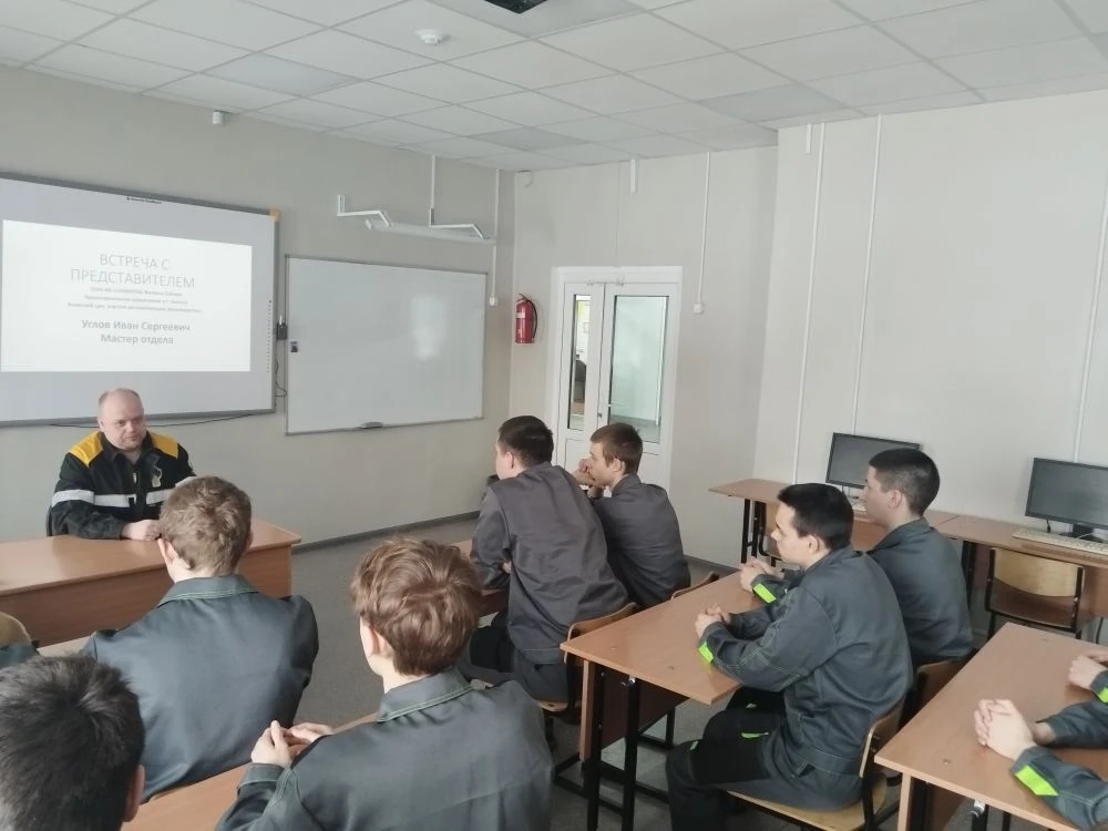 группа молодых людей сидит за столами в учебном классе. Ведущий, также в черно-желтой куртке, находится впереди и объясняет материал. На стене проектора или белой доске отображается текст, вероятно, с информацией или презентацией. Молодые люди выглядят заинтересованными и внимательными к лекции. Комната оформлена просто, с несколькими компьютерами на столах, создавая сосредоточенную атмосферу обучения.