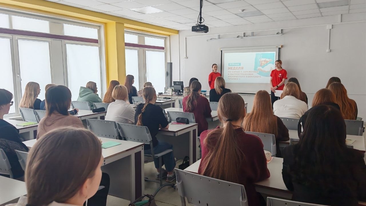 изображен класс, в котором проходят занятия. Слева стоят двое выступающих в красных футболках и демонстрируют презентацию на экране. В аудитории присутствует много студентов, которые сидят за партами и внимательно слушают. На стенах комнаты есть окна с естественным освещением и проектор, установленный на потолке. Обстановка выглядит учебной и сосредоточенной.