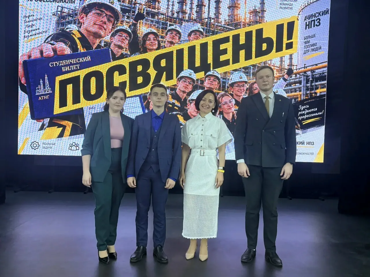 Четверо ведущих (две женщины и двое мужчин) стоят в ряд на сцене перед большим экраном. Слева — женщина в темно-зеленом брючном костюме и розовой футболке. Рядом с ней — молодой человек в темно-синем тройном костюме и ярко-синей рубашке. В центре — женщина в белом фактурном платье миди с поясом и бежевых туфлях. Справа — мужчина в темном двубортном костюме с галстуком. На заднем плане на экране крупная желтая надпись «ПОСВЯЩЕНЫ!», коллаж из портретов студентов в касках, логотип Ачинского НПЗ и макет синего студенческого билета с логотипом АТНГ.