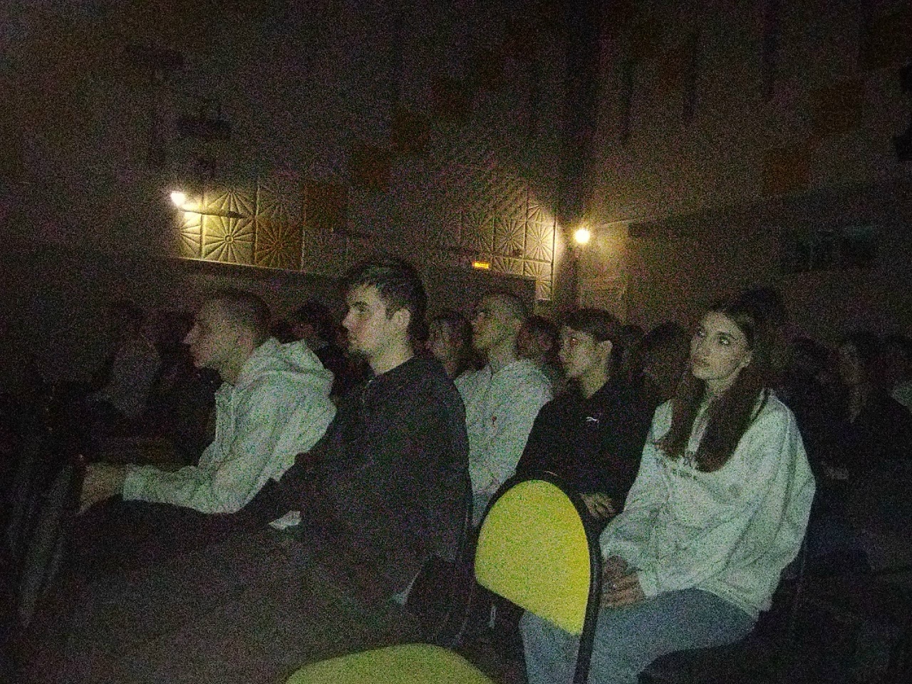 На фотографии изображены люди, сидящие в аудитории. Все они внимательно смотрят вперёд, возможно, на экран или на выступающего. В комнате приглушённый свет, создающий атмосферу сосредоточенности. На переднем плане видно несколько человек в светлой и тёмной одежде. Некоторые сидят с поднятыми плечами или скрестив руками. В общем, сцена выглядит довольно камерно и сосредоточенно, передавая ощущение совместного просмотра или обсуждения.