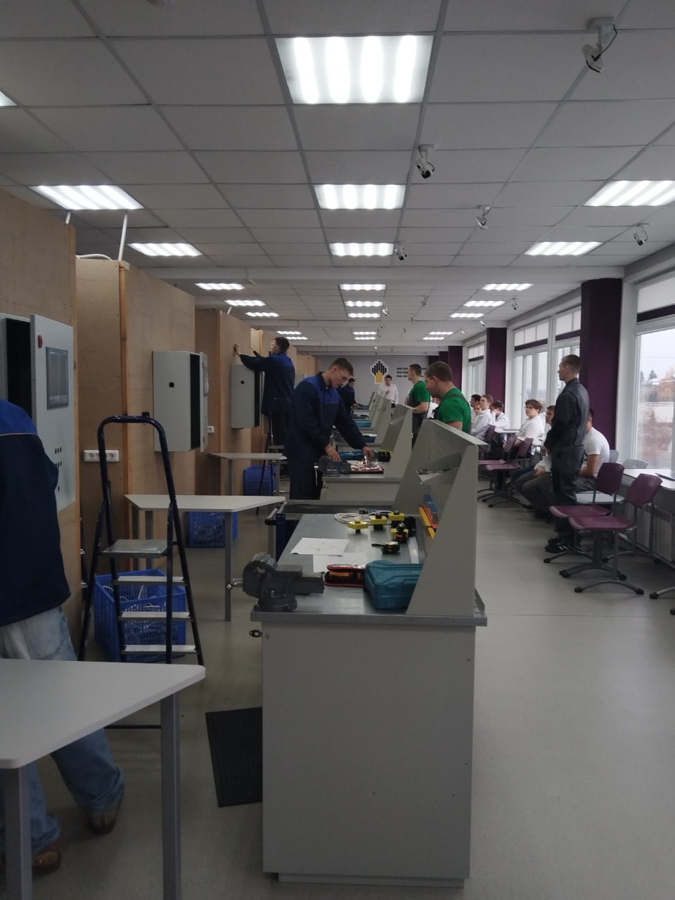 На этом фото показано светлое помещение, в котором несколько людей работают за столами. Столы организованы для выполнения практических заданий, на них находятся разные инструменты и детали. Видны стенды на заднем плане, где также работают студенты, и две большие тумбы с оборудованием.