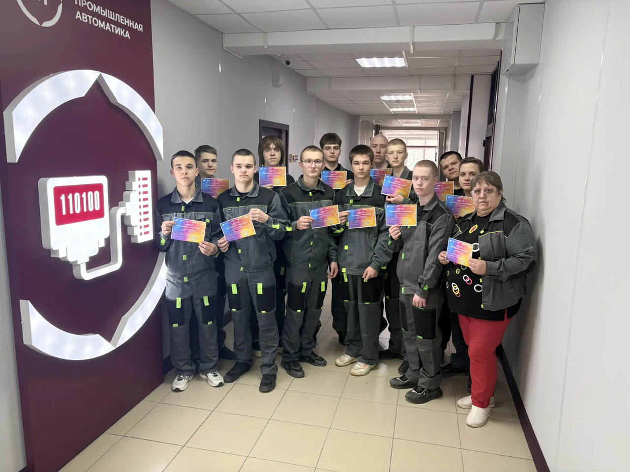 Группа людей в коридоре: около пятнадцати молодых мужчин в тёмно‑серой рабочей форме и одна женщина справа в повседневной одежде. Все стоят в несколько рядов, держат в руках яркие цветные сертификаты (градиентные карточки), смотрят в камеру. Слева на стене крупный круглый знак с пиктограммой и цифрами «110100». Освещение офисное, плиточный пол tiled пол и светло‑серые стены.