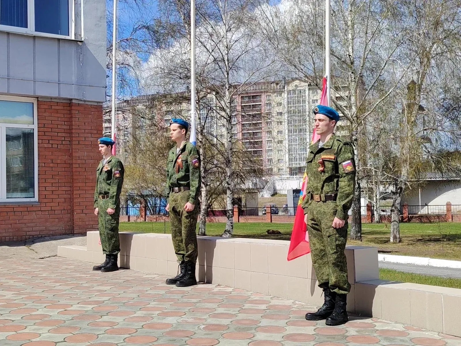 На этом изображении три военнослужащих, стоящих в ряд. Они одеты в военную форму и имеют голубые береты на головах. Сзади них виден флаг и элементы инфраструктуры, такие как здания и деревья. Атмосфера выглядит торжественной, на улице ясный день с облаками на небе.