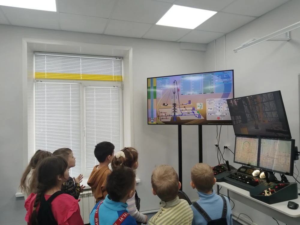 фотография показывает группу детей, которые наблюдают за экраном. На экране представлены данные или информация в формате графиков и чисел. Дети обращают внимание на экран, некоторые улыбаются или выражают интерес. Они также одеты в яркие одежды и комбинезоны. Освещение в помещении яркое и хорошо распределенное.