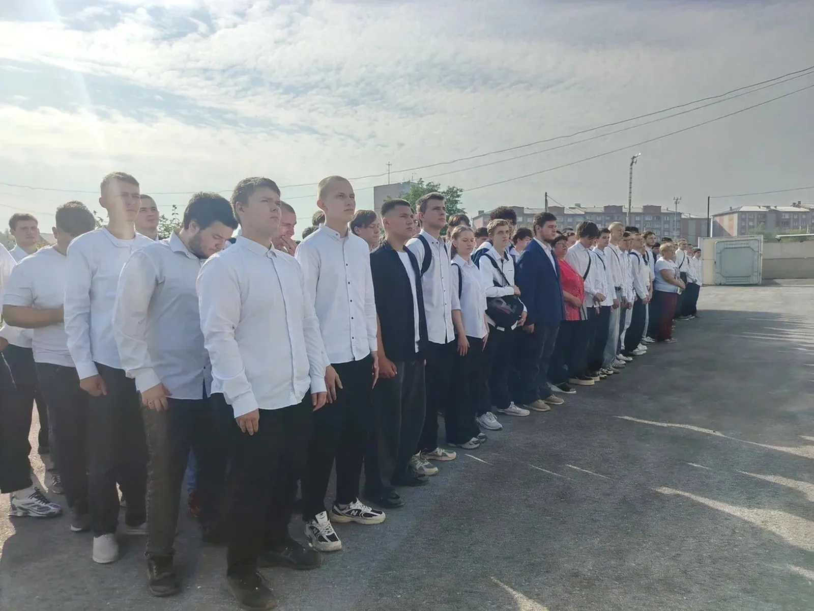 Большая группа школьников и студентов в парадной форме (белые рубашки, темные брюки и юбки) стоят ровными шеренгами на асфальтированном плацу под открытым небом во время торжественной линейки. Позади них находятся многоэтажные жилые дома и линии электропередач.