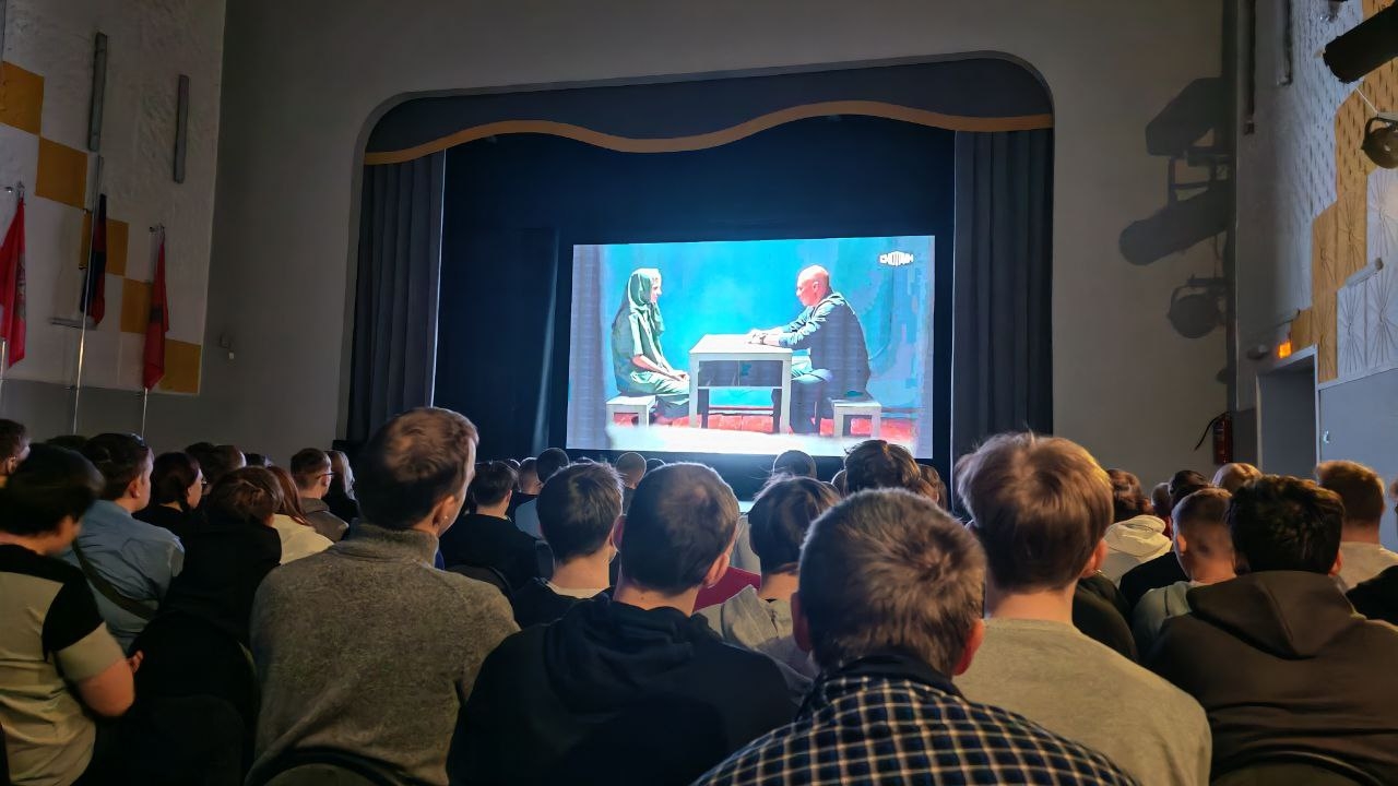 На фото изображена сцена из кинозала, где аудитория внимательно смотрит на экран. На экране также представлены два человека, похожие на тех, что на постере. Аудитория состоит в основном из молодежи, которая сидит спиной к камере, и их лица не видно. Сцена освещена мягким светом, создающим атмосферу ожидания.