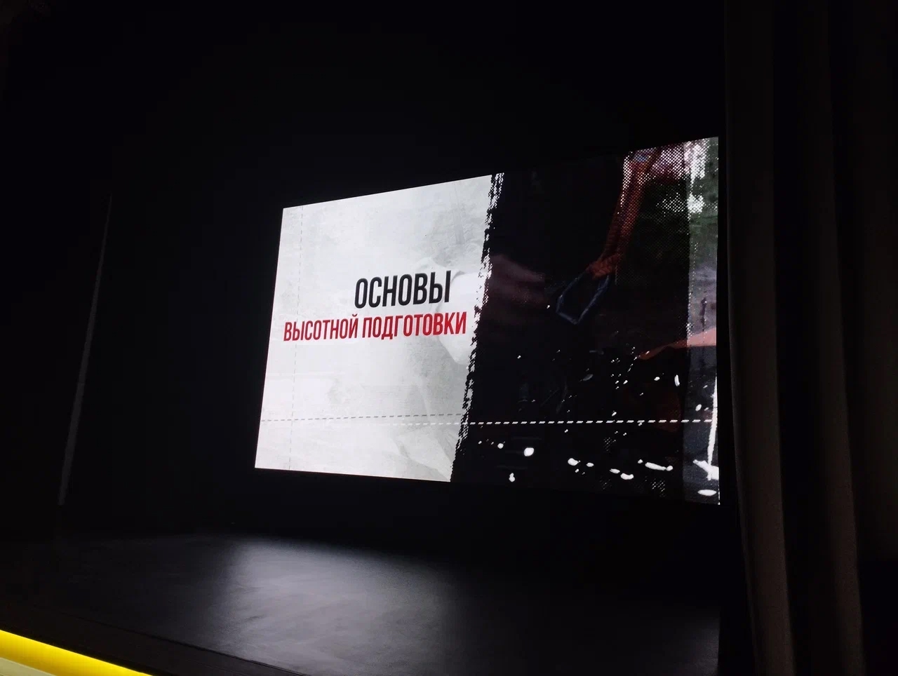 На экране проектора отображается текст "Основы высотной подготовки". Фон черный, а текст белый и красный, что делает его хорошо читаемым. Сцена выглядит подготовленной для презентации или лекции, с минималистичным оформлением.