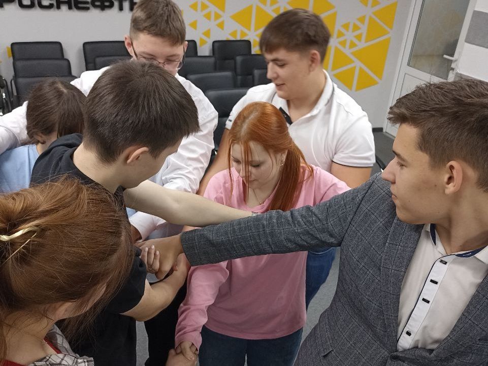 На изображении группа молодых людей, занимающихся командной деятельностью. Несколько мужчин и женщин стоят в кругу, скрестив руки. В центре находится девушка с длинными рыжими волосами, она явно вовлечена в процесс. На заднем плане видны стулья и светлая стена с логотипом. Атмосфера кажется динамичной, есть ощущение партнёрства и сотрудничества.