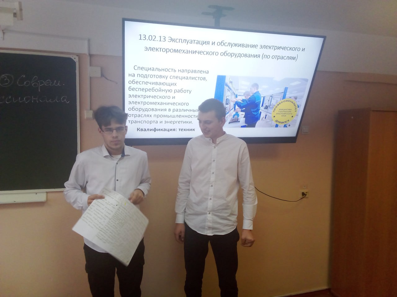 На кадре видно двоих мужчин, стоящих перед экраном с презентацией: Один из них держит лист бумаги, возможно, с заметками или планом. Второй мужчина активно объясняет что-то, указывая на экран, на котором отображены учебные материалы, связанные с электротехникой. Фоновый элемент включает домашних библиотеки и доску, что подчеркивает учебную атмосферу.