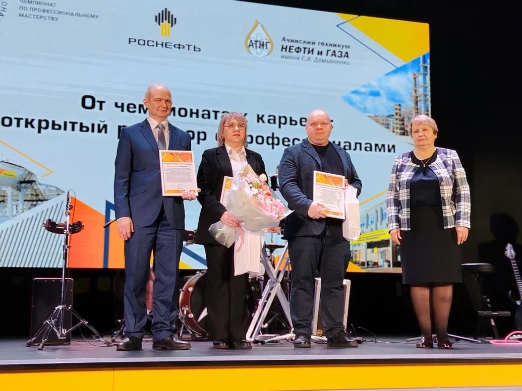 На сцене стоят четыре человека, все улыбаются. Слева направо: мужчина в строгом костюме с галстуком, женщина в деловом костюме с цветами, мужчина в очках с документами, и женщина в светлом платье. За ними большой экран с логотипом и текстом, посвященным профессиональному мастерству.