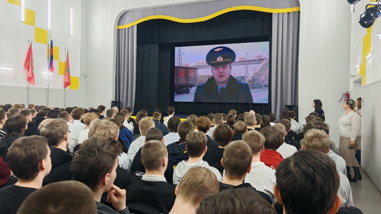 руппа молодежи внимательно смотрит на экране, где транслируется видеозапись человека в форменной одежде, вероятно, офицера полиции или представителя правоохранительных органов. Женщина на сцене продолжает вести мероприятие. Зал заполнен светлыми головами, а на заднем плане можно увидеть флаги и яркую архитектуру.