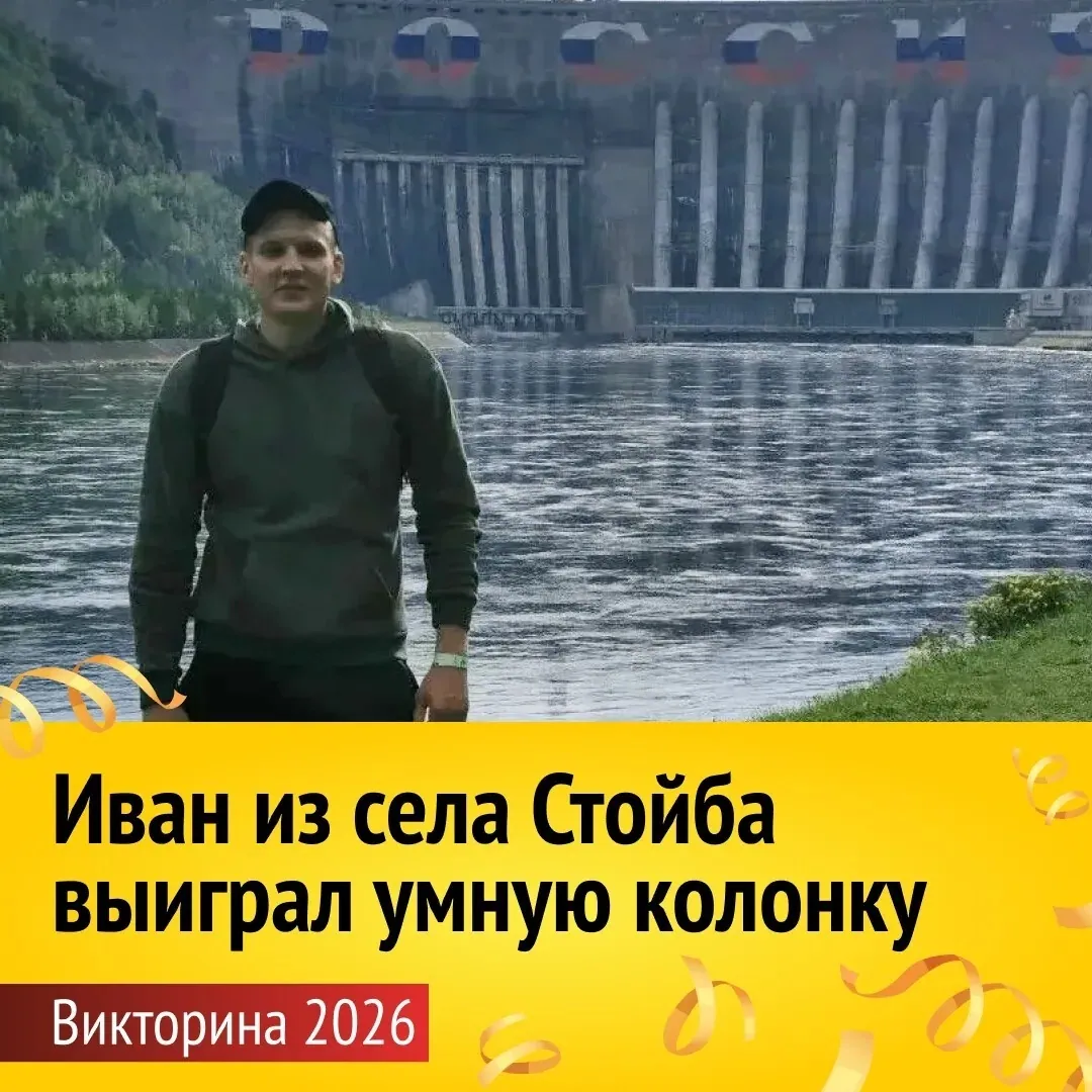 Мужчина в темной кепке и зеленом худи стоит на берегу реки. На заднем плане — бетонная плотина с флагами России. Внизу надпись: «Иван из села Стойба выиграл умную колонку. Викторина 2026».