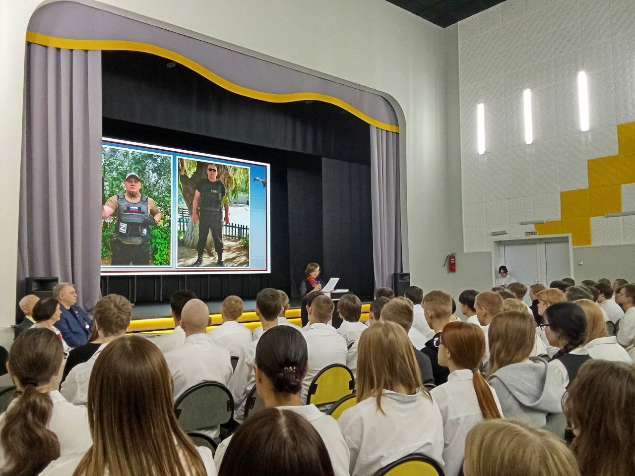 показана сцена в большом зале. На сцене стоят два экрана, на которых изображены два человека. Слева – мужчина в тактическом снаряжении с нагрудными знаками, а справа – женщина, одетая в формальные одежды, с темными очками и короткими волосами. В зале сидит много людей, в основном молодежь в белых рубашках. Они обращают внимание на выступление человека на сцене, который, вероятно, делает доклад или презентацию.