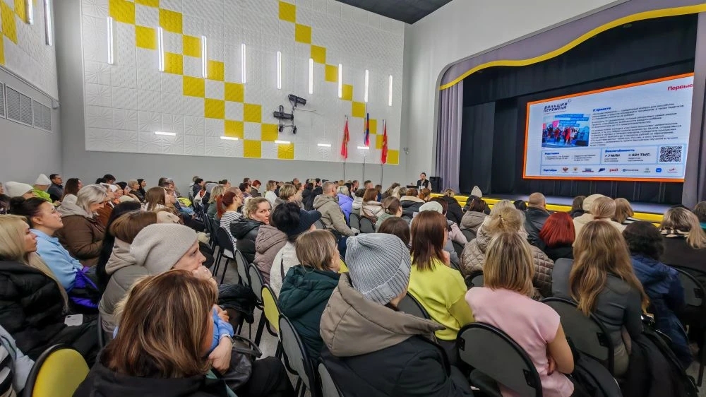 фото показан просторный зал, заполненный людьми. В центре находится большая проекционная экран, на котором отображается информация. Существует группа слушателей, расположенная в зале; большинство из них одеты в светлую или нейтральную одежду. На заднем фоне видны несколько флагов, а стены оформлены в желто-белых тонах.