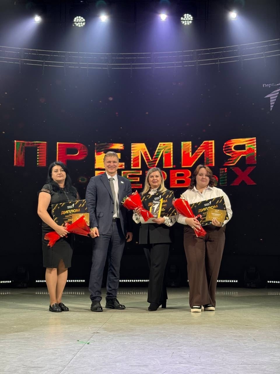 На сцене находятся три женщины и мужчина. Женщины стоят слева и справа, одна из них в черном костюме, а другая — в светлом. Мужчина в сером костюме находится в центре и держит диплом, а все трое женщин улыбаются и держат цветы. На заднем плане снова виден экран с ярким названием "Премия Первых".