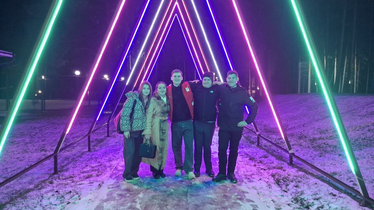 На этом фото группа из пятерых человек стоит на улице в окружении ярких неоновых огней. Светящиеся треугольные конструкции на заднем плане создают эффект арки и окрашены в различные яркие цвета: от зеленого до фиолетового. Снег покрывает землю, и вечернее освещение создает атмосферу праздника. Люди стоят с улыбками и уверенными позами: мужчина в красной куртке, рядом с ним девушка в длинном бежевом пальто, затем несколько мужчин, один из которых одет в черную куртку. Все выглядят расслабленно и счастливо, как будто собираются отпраздновать что-то важное.