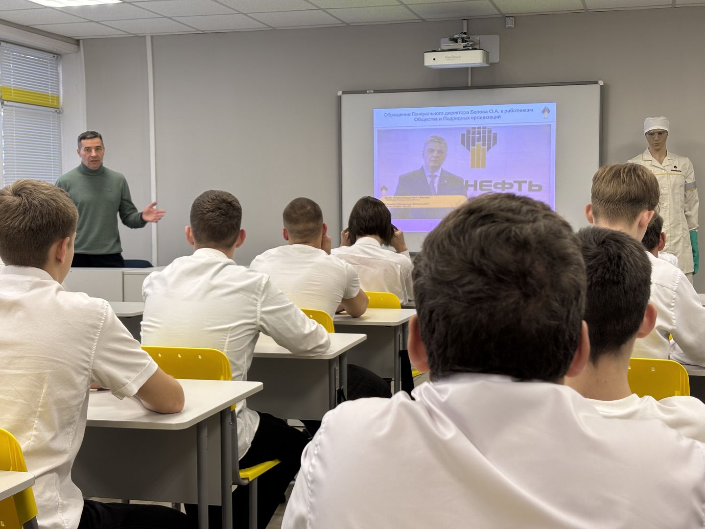 На первом изображении мы наблюдаем класс, в котором сидят студенты, смотрящие в сторону преподавателя или экрана. Перед ними расположен большой экран с презентацией, на которой виден текст и изображение, возможно, связанное с темой урока (в данном случае, о нефти). Преподаватель стоит слева от экрана, обращаясь к ученикам. Он одет в тёмный свитер и выглядит уверенно. Студенты, в белых рубашках, внимательны и сосредоточены на материале. Позади него находится манекен в рабочей одежде, что может указывать на направление специальности, изучаемой в классе.
