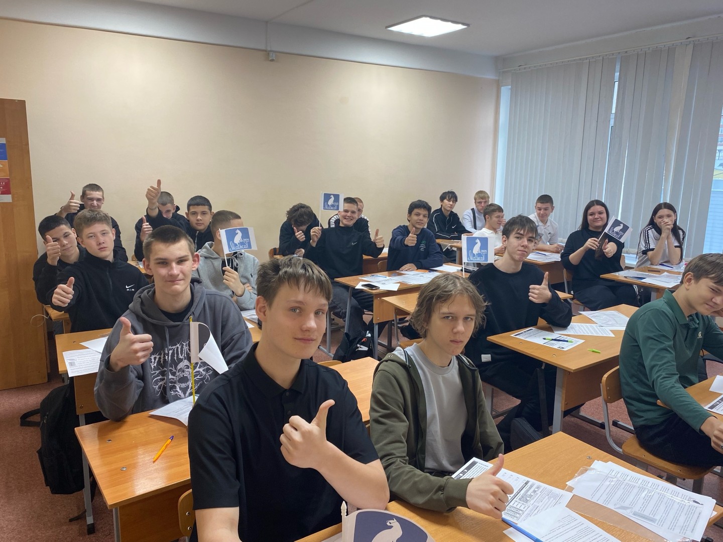 На данном фото представлена ещё одна группа из пятнадцати учеников, сидящих за столами в светлом классе. Все они демонстрируют жесты "ок", что сигнализирует о том, что они довольны чем-то. Как и в первой фотографии, на столах лежат рабочие листы, а в руках у многих флажки с изображением, возможно, связанным с проектом или мероприятием. Виден хороший доступ к естественному освещению через окна.