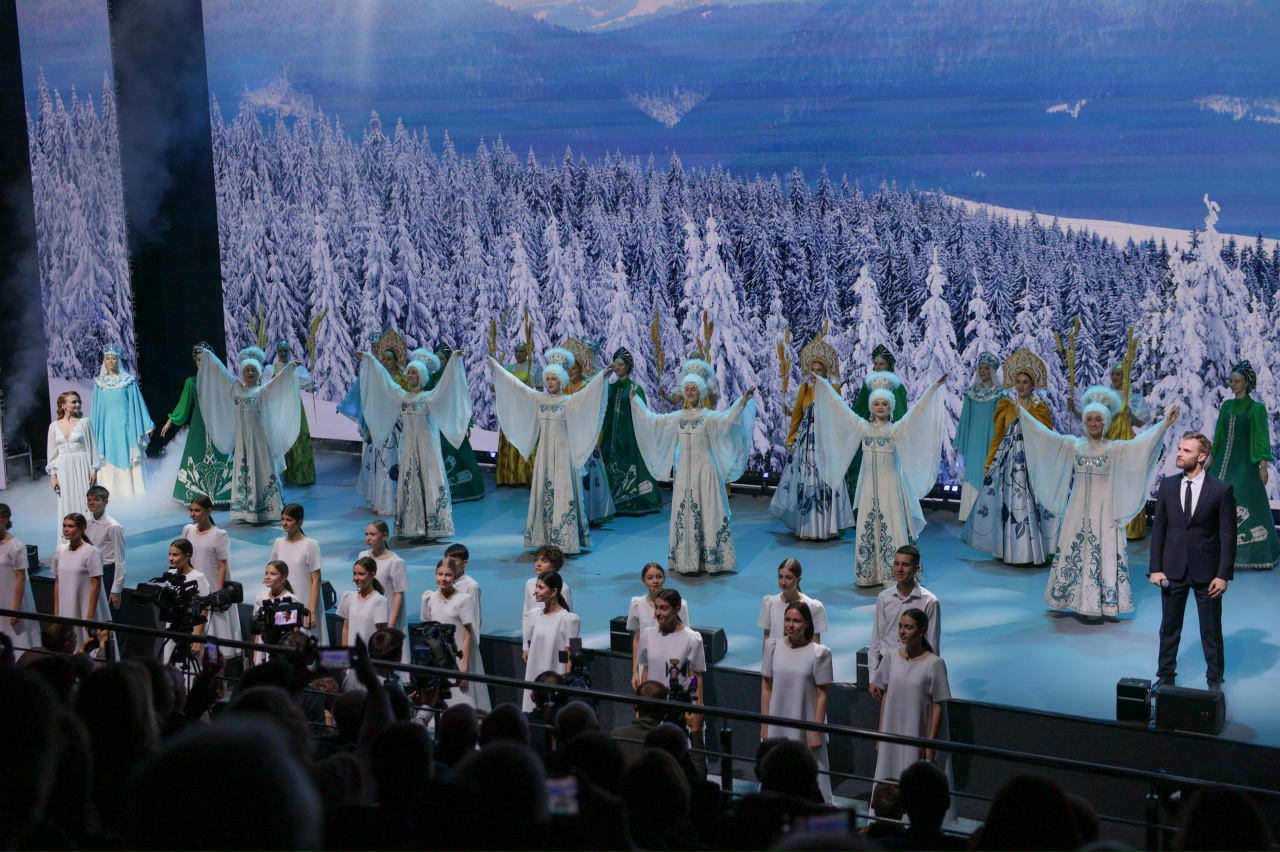 На этом фото сцена заполнена множеством артистов, одетых в белые и цветные костюмы. Они находятся на фоне красивого зимнего пейзажа с заснеженными деревьями и горами. Артисты выполняют хоровое пение и движения руками, создавая эффект гармонии и единства. В аудитории видны зрители, которые внимательно наблюдают за выступлением, создавая атмосферу праздника и зрительского восторга.