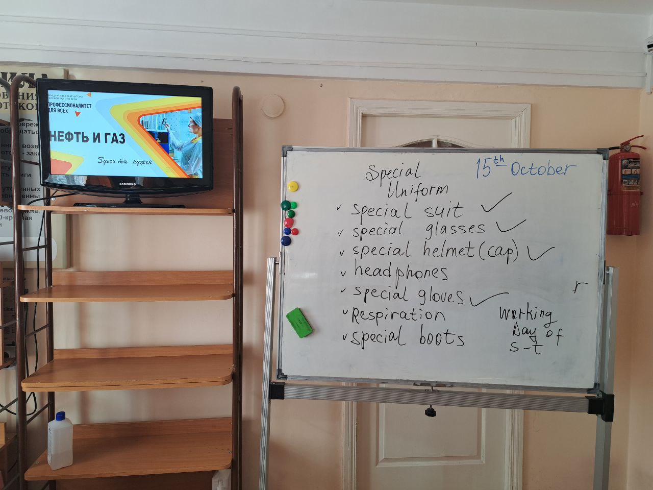 На фото видно класс, где на белой доске написаны записи о специальной униформе, требуемой для работы, перечисленные пункты включают специальный костюм, очки и каску. На стене висит телевизор, на экране которого идет трансляция с изображением. Рядом с доской стоит стеллаж.