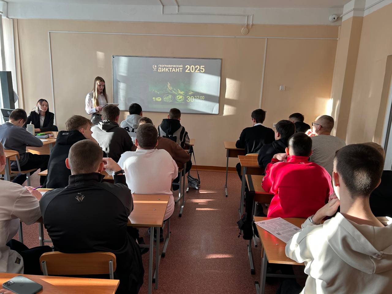 На фото показана классная комната. В центре внимание сосредоточено на экране, на котором написано "Итог 2025". На переднем плане, справа, стоит преподавательница, которая ведет урок. За ней расположены ученики, сидящие за партами. Они выглядят сосредоточенно и внимательно смотрят на экран.