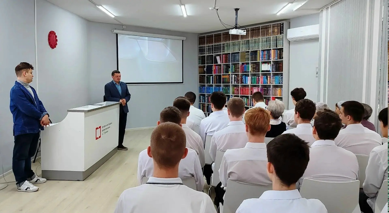 сцена с презентацией в том же помещении. Большая часть внимания сосредоточена на двух мужчинах: один из них стоит у стола, а другой — рядом с ним, чуть правее. Первый говорит к аудитории, сидящей перед ним. На стене проекция слайдов, а за спинами сидящих виден стеллаж с книгами в ярких обложках. Атмосфера более формальная, с акцентом на образовательный процесс.