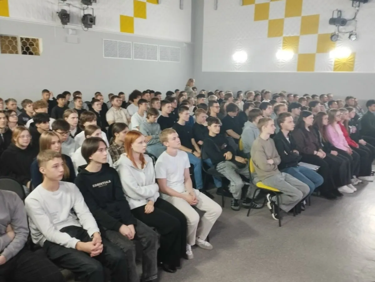 Большая группа подростков и молодых людей сидит рядами в светлом зале с желто-белыми стенами. Ребята одеты в повседневную одежду и внимательно смотрят вперед, слушая выступающего.