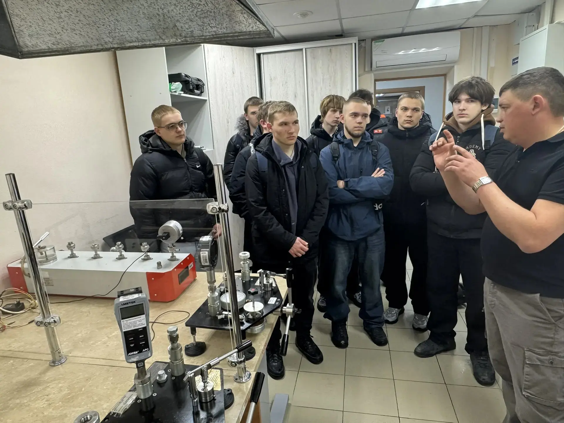 На этом изображении группа из шести человек, стоящих перед столом с научным оборудованием. Инструктор объясняет что-то, показывая детали на приборе, в то время как остальные участники внимательно его слушают и смотрят. В комнате много различного оборудования, некоторые приборы находятся под защитным стеклом.