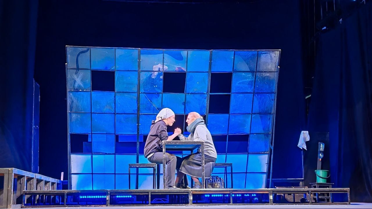 На сцене театра два актёра сидят за столом и, похоже, ведут эмоциональное соревнование или дискуссию. Они оба в тёмных одеждах и с платками на голове, сидят напротив друг друга и внимательно смотрят друг на друга. Фон состоит из больших стеклянных панелей, возможно, с синеватыми оттенками, создающих атмосферу напряжённости или драмы. Сцена освещена синим и белым светом, что усиливает эффект происходящего.