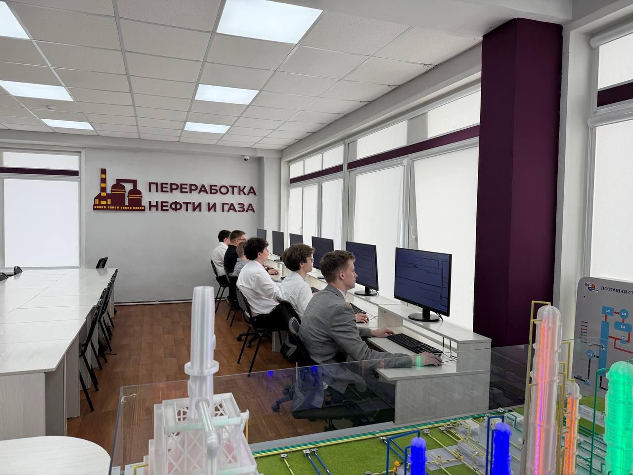На фото представлено более просторное помещение, где за компьютерами сидят несколько сотрудников в белых рубашках. На заднем плане расположен большой макет завода по переработке нефти. Стена оформлена в темных тонах с надписью «Переработка нефти и газа». В интерьере много света благодаря большим окнам, оформленным белыми шторами.