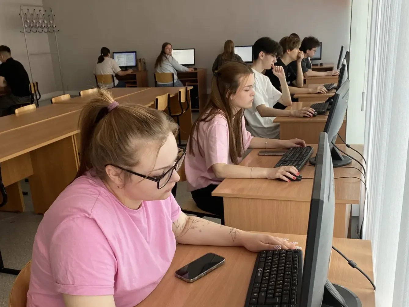 Фотография просторного учебного компьютерного класса. На переднем плане две девушки сидят за деревянными партами. Девушка в розовой футболке и очках сосредоточенно смотрит в монитор компьютера, её правая рука лежит на мышке, рядом на столе находится смартфон. В глубине комнаты за аналогичными столами сидят другие студенты. Помещение светлое, с окнами справа, у стены слева висит металлическая вешалка для одежды.