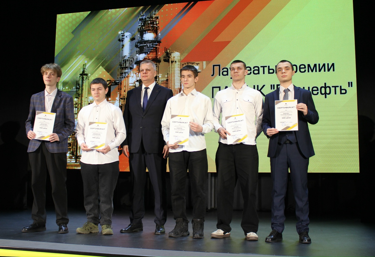 изображена группа из пяти молодых мужчин, стоящих на сцене. Они одеты в светлые рубашки и один из них в темном пиджаке. На фоне яркий экран с графическими элементами и написано: "Лауреаты премии ПАО "Нефть"". У всех лауреатов в руках сертификаты, их выражения лиц показывают гордость и радость. На сцене также присутствует мужчина в строгом костюме, вероятно, передающий награды.