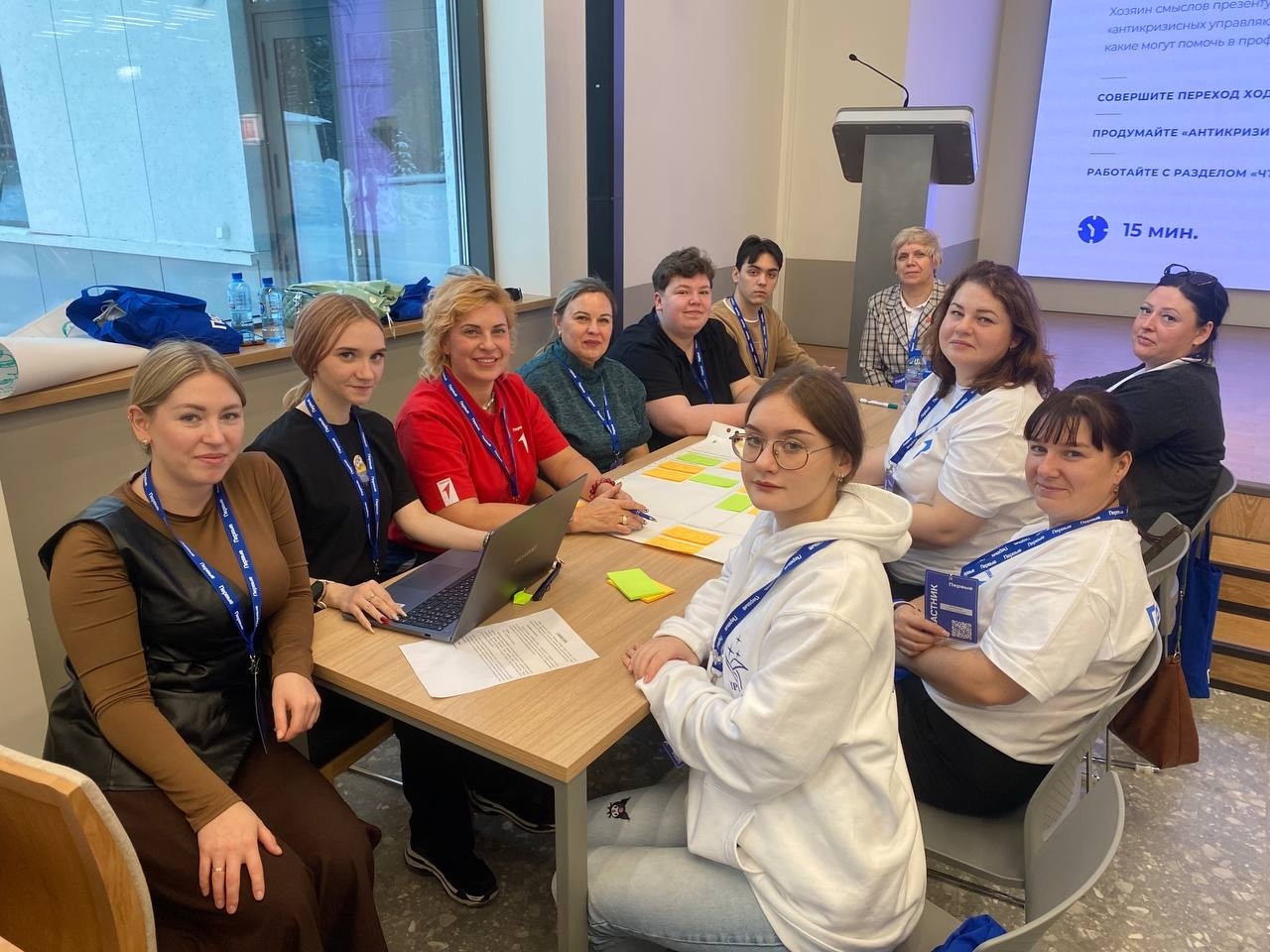 На изображении группа из десяти человек, сидящих за столом. Женщины разных возрастов и внешности, некоторые из них носят униформу или логотипы мероприятия. На столе виден ноутбук и листы бумаги с заметками. Атмосфера дружелюбная и рабочая, все активно общаются и обсуждают что-то важное.